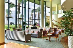 Un hall d’entrée spacieux, doté d’une grande fenêtre offrant une vue sur des palmiers et un jardin. On y trouve plusieurs coins salon avec des canapés et des fauteuils.