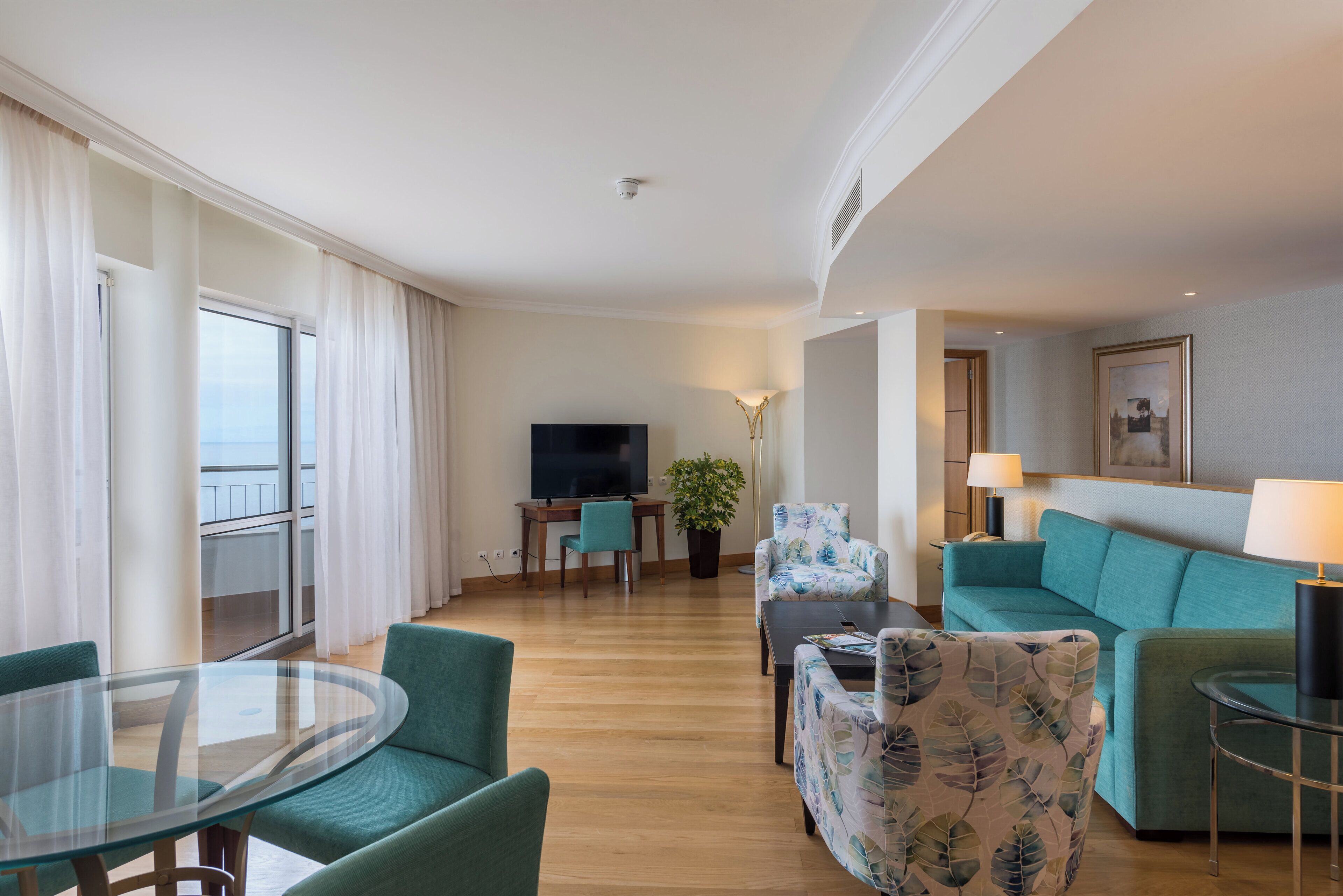 Ein modernes Hotelzimmer mit Balkon, einem Flachbildfernseher, einem Schreibtisch mit Stuhl, einem Sofa, einem gemusterten Sessel, einem Couchtisch und einer Lampe.