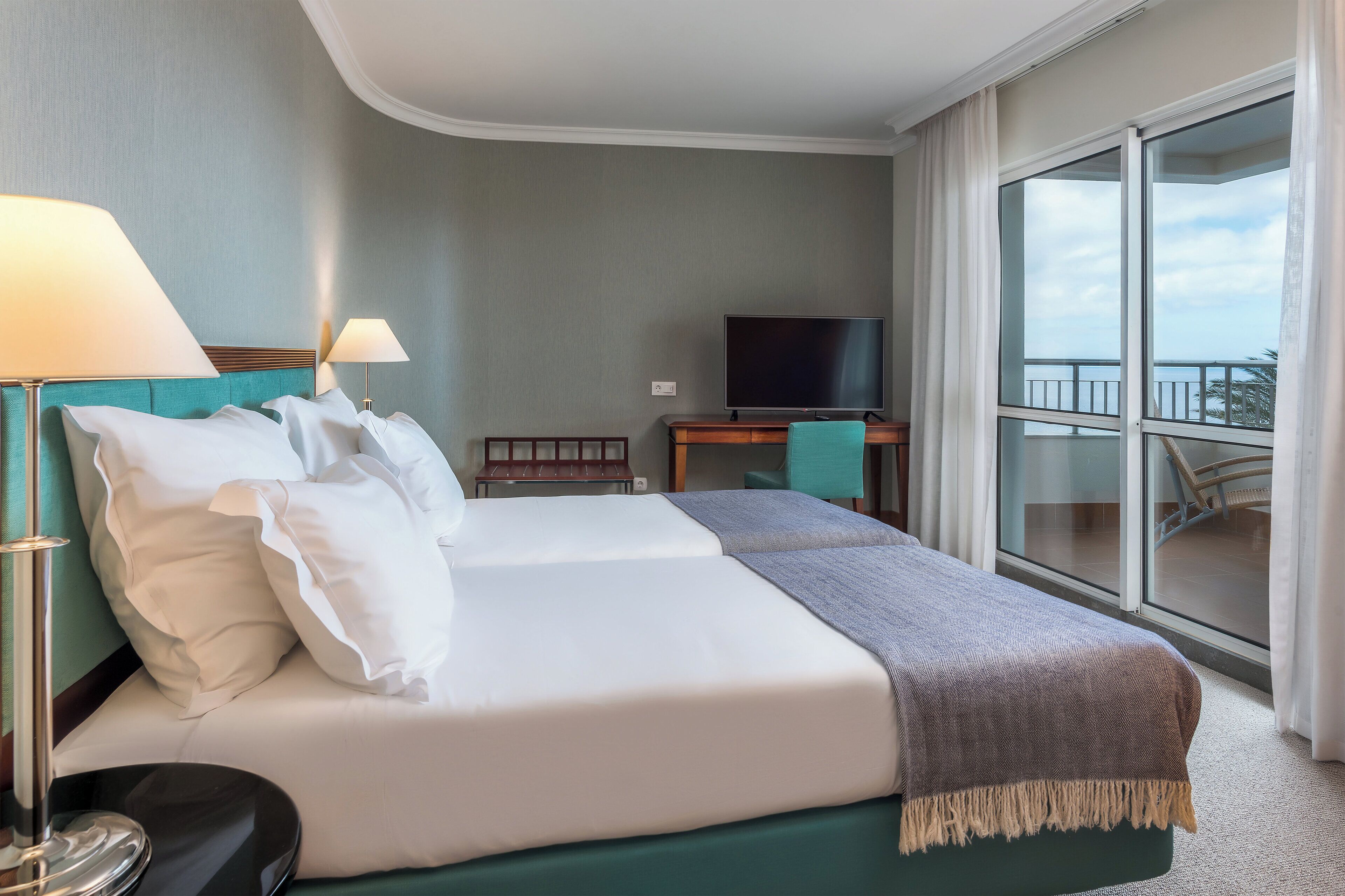 A hotel room with a bed, two bedside lamps, a television, a desk, and a view of the balcony.
