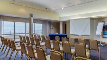 Un salón de conferencias con filas de sillas, una pantalla para proyector y una pizarra para presentaciones.