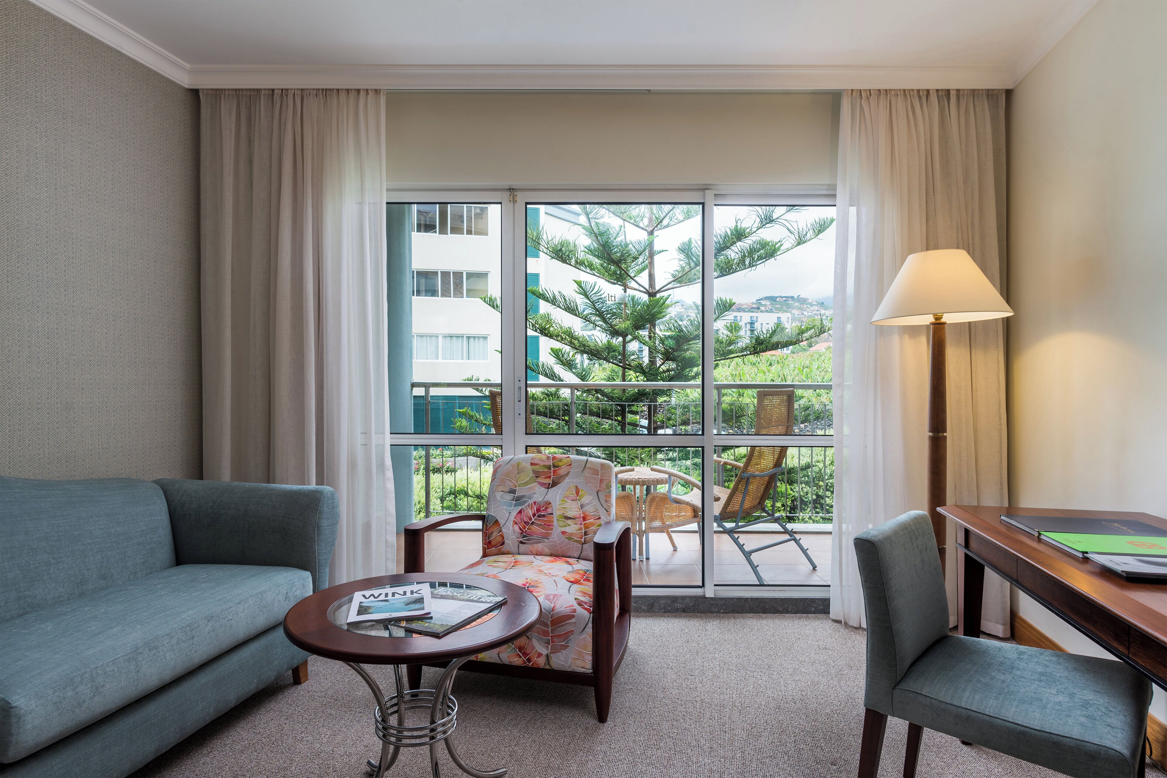 Habitación de hotel con sofá, una silla, una mesita, lámpara y vistas a zonas verdes y edificios a través del ventanal.