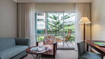 Ein Hotelzimmer mit Sofa, Sessel, kleinem Tisch, Lampe und Blick auf Grünflächen und Gebäude durch das Fenster.