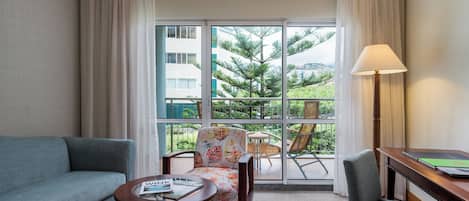 A hotel room with a sofa, a chair, a small table, a lamp, and a view of greenery and buildings through the window.