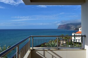 Un balcone con ringhiera in metallo che si affaccia su una vista costiera, con cielo sereno e una montagna in lontananza.