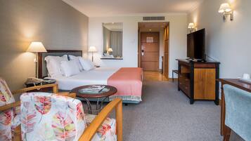A hotel room with a bed, bedside lamps, a desk with a television, a chair, and a small table with a magazine.