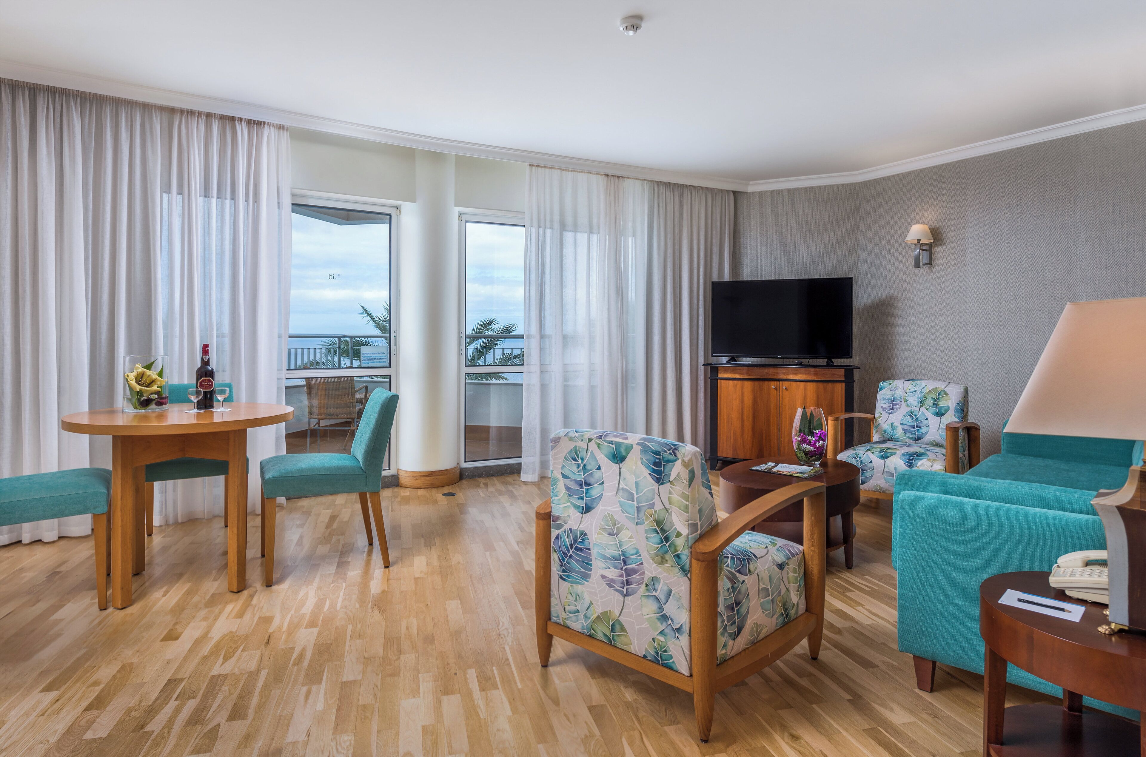 A hotel room with a wooden floor, a dining table with chairs, a sofa, a television, and a balcony with curtains.