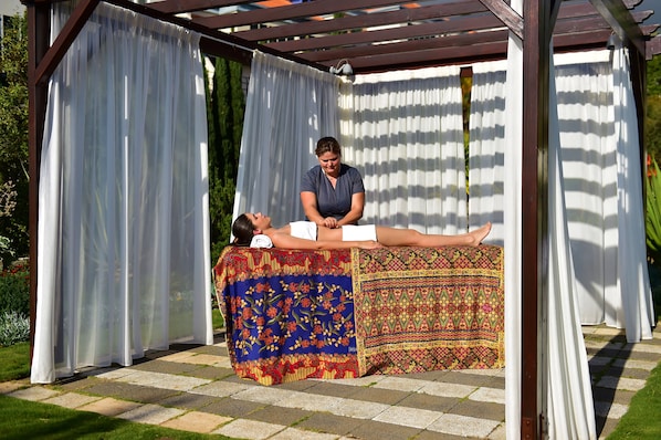 Une personne allongée sur une table de massage à l’extérieur, sous une pergola surmontée de rideaux blancs.