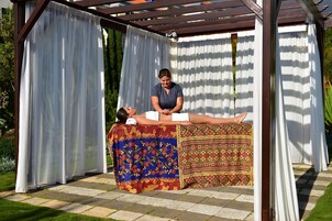 Una persona recostada en una camilla de masajes al aire libre, bajo una pérgola con cortinas blancas.