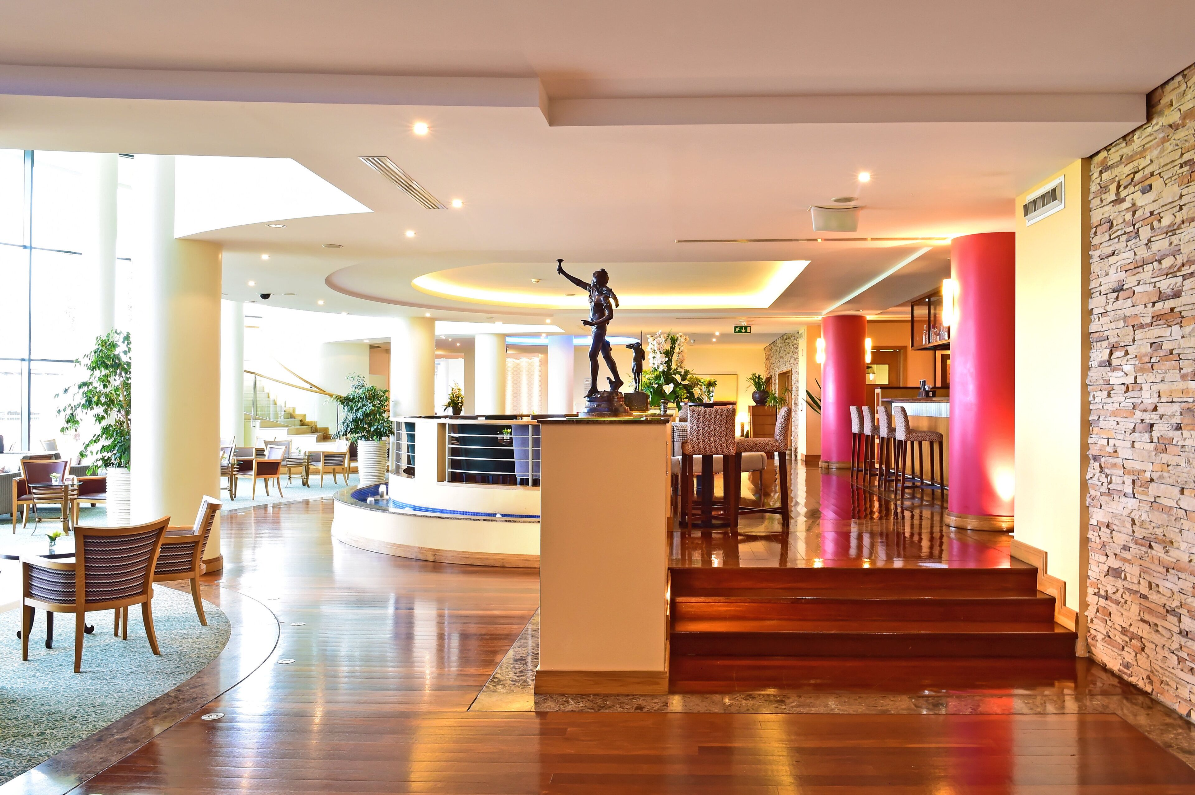 A hotel lobby with a central reception desk, seating area, and a staircase leading to upper floors.
