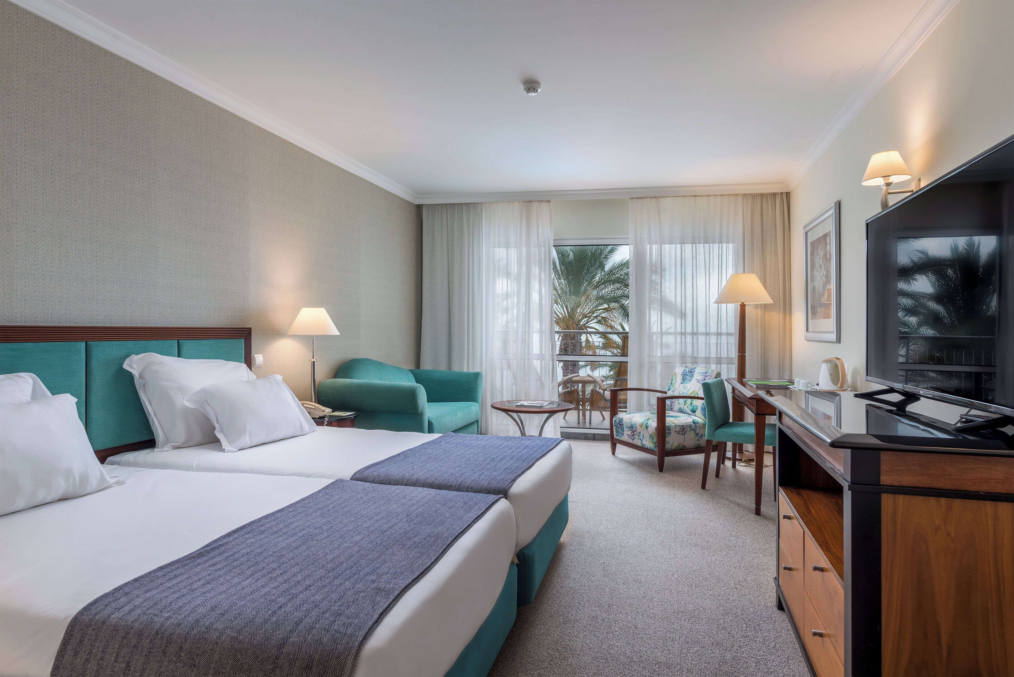 A hotel room with a bed, two chairs, a desk, a television, and a window with curtains.