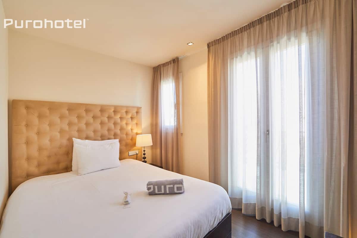 Een hotelkamer met een gestuikt hoofdeinde, een bed met witte lakens, een bedlamp en transparante gordijnen.