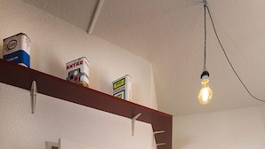 A hallway with a white door, a shelf with paint cans, a hanging light bulb, a mounted TV, and a green trash bin. - Artyster Clermont-Ferrand (Clermont-Ferrand)
