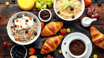 Mesa de café da manhã com tigelas de aveia, iogurte e granola, acompanhada de croissants, uvas, morangos e café.