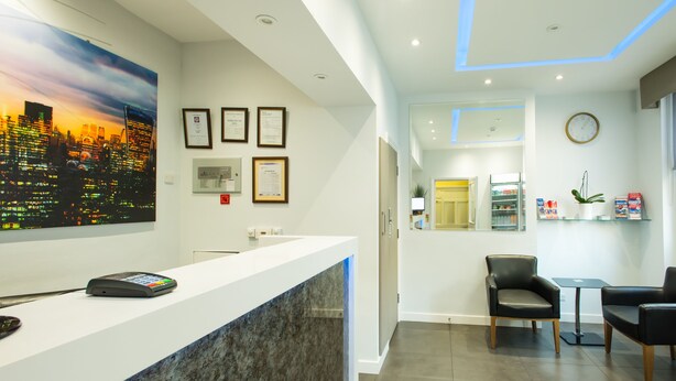 A modern reception area with a white counter, a cityscape painting, framed documents, a clock, and a small seating area. - Marble Arch Inn Hotel (London)
