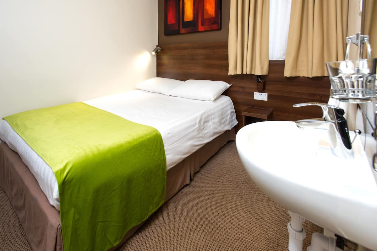 A hotel room with a bed, a green bedspread, a white pillow, a wooden headboard, a beige carpet, a white sink, and a mirror.