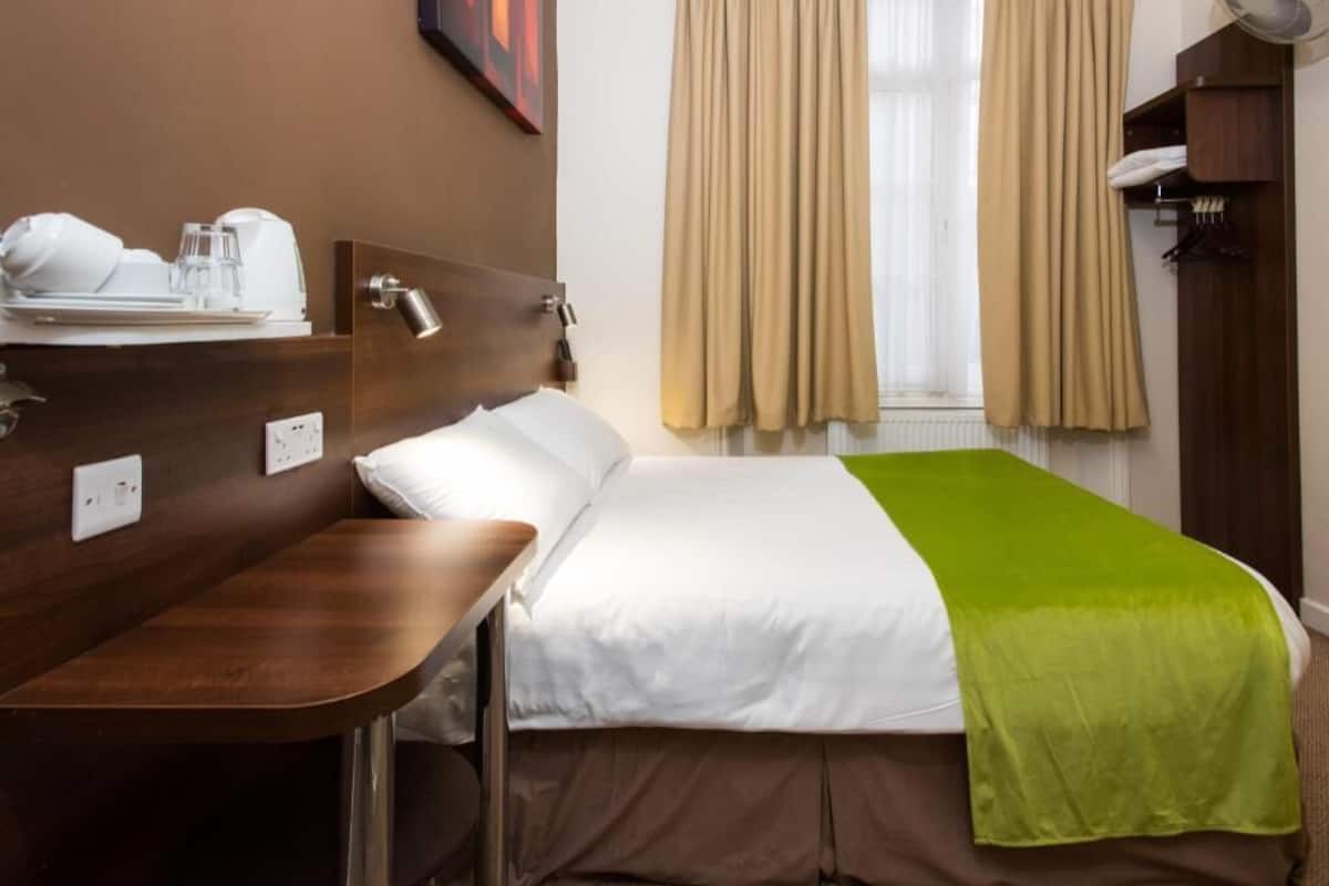 A hotel room with a bed, a bedside table, a lamp, a window with curtains, and a wall-mounted shelf.