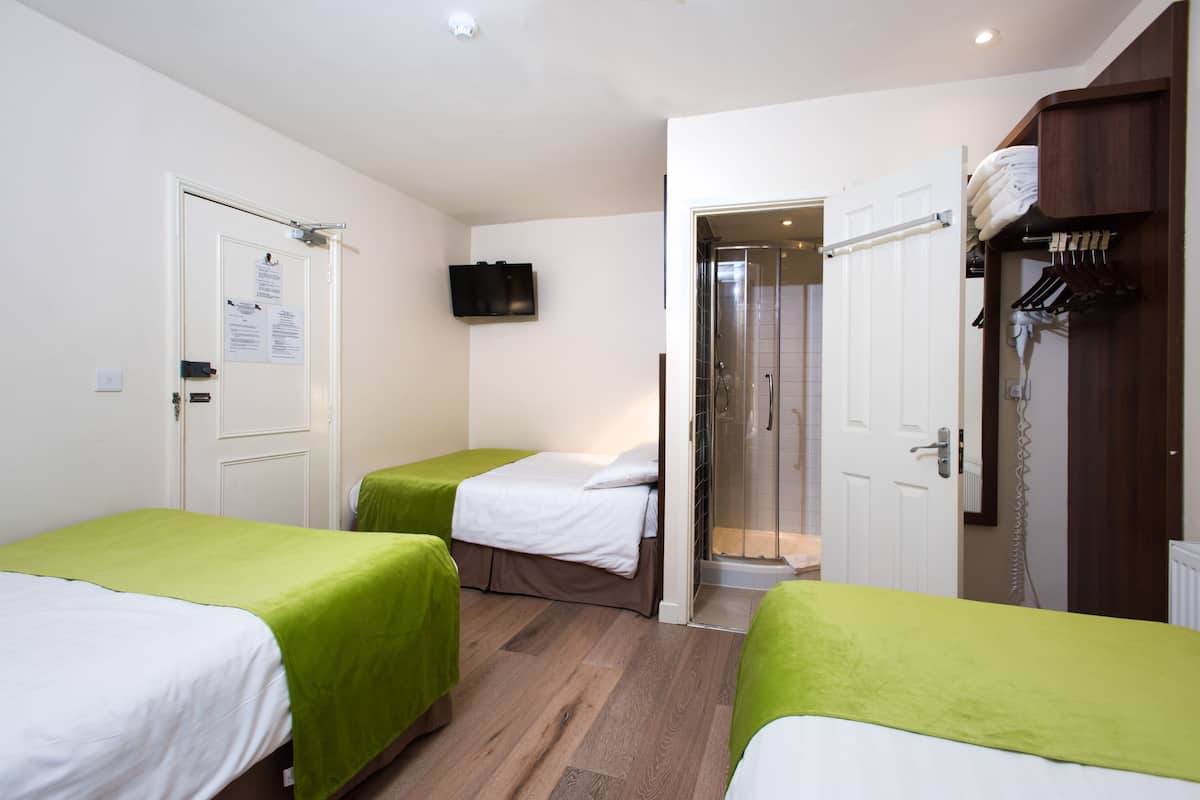 A hotel room with two single beds, a shower area, a television mounted on the wall, and a wardrobe with hanging clothes.