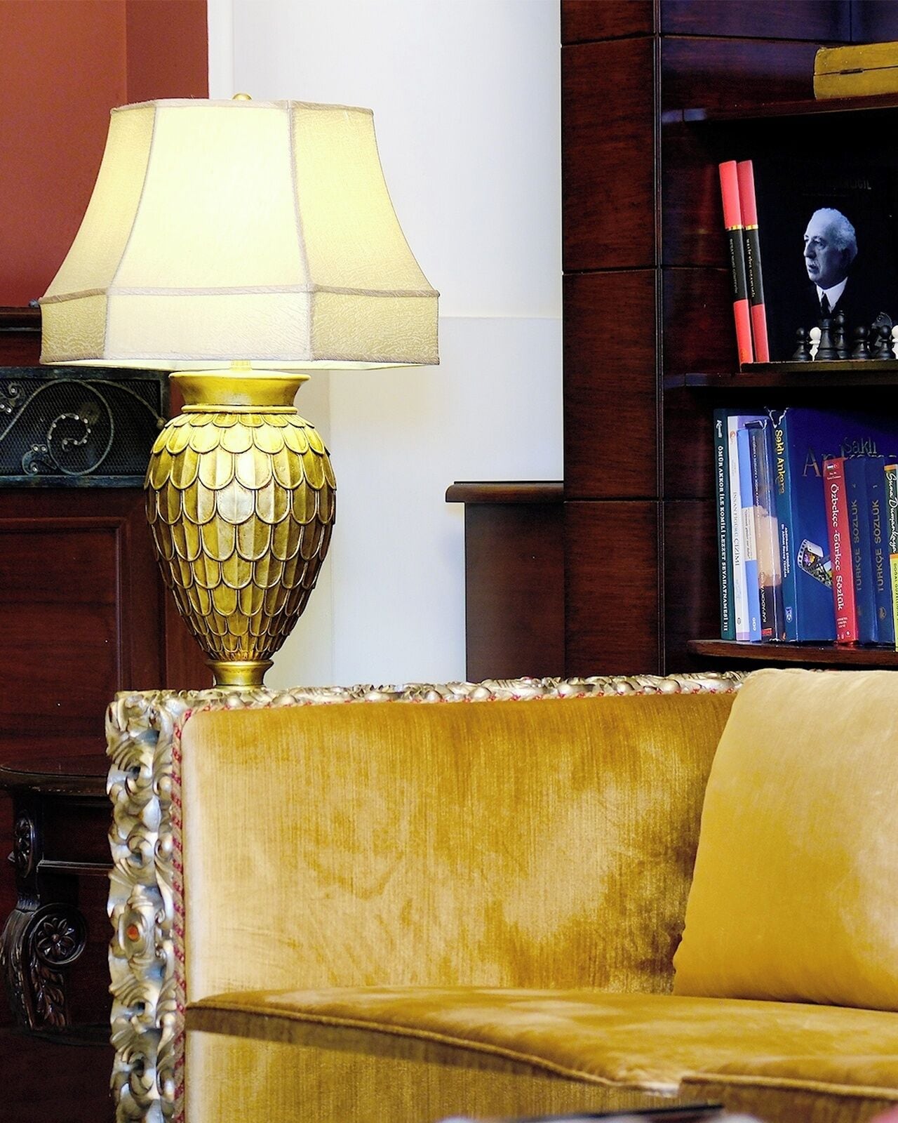 A room with a gold-colored sofa, a decorative lamp with a gold base, and a bookshelf with books and a chess set.