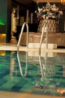 Zona de piscina con piso de vidrio transparente, un sofá color crema y un arreglo floral sobre un pedestal.
