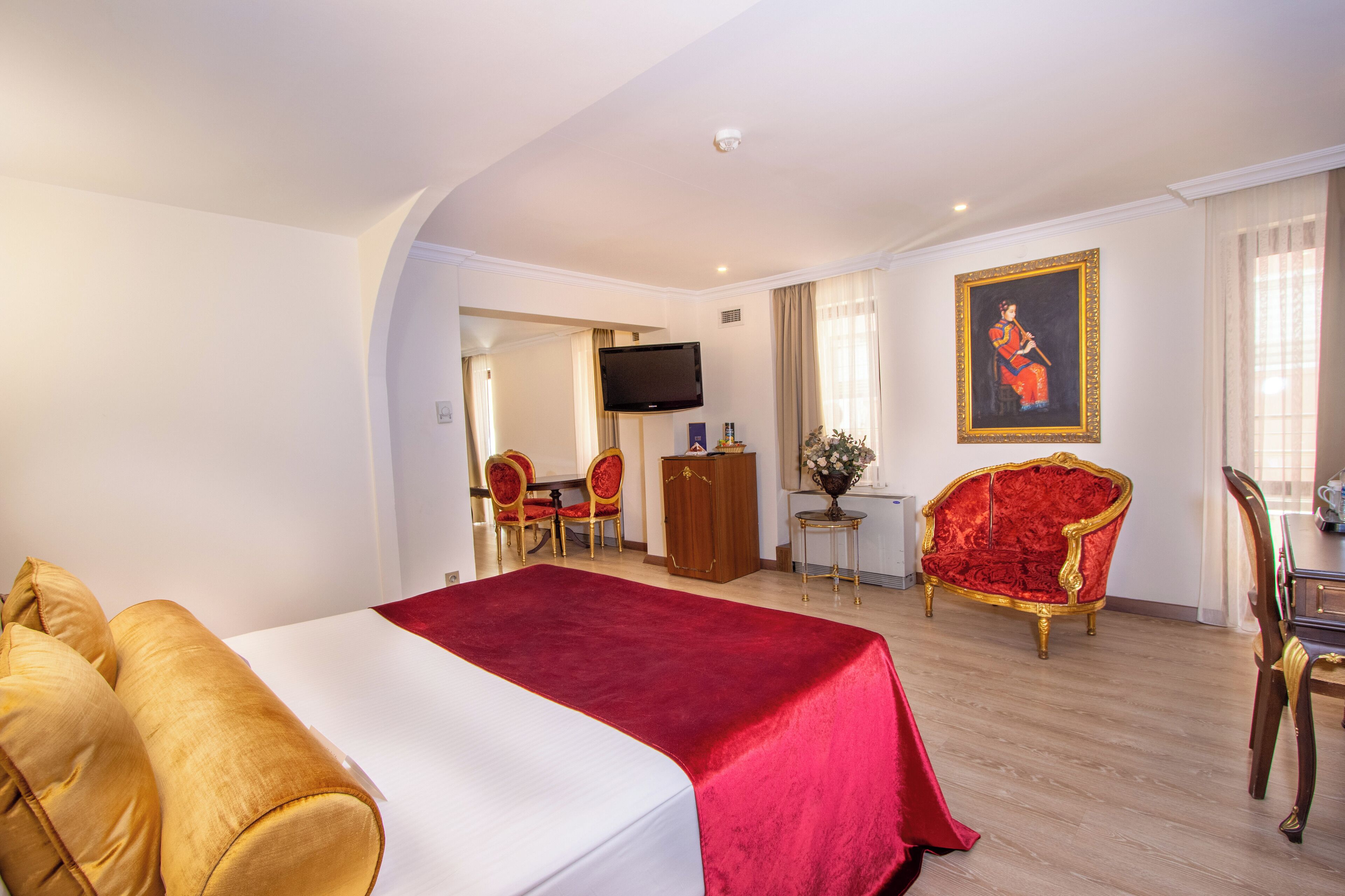 A hotel room with a large bed, a red upholstered chair, a wooden desk, a television, and a painting on the wall.