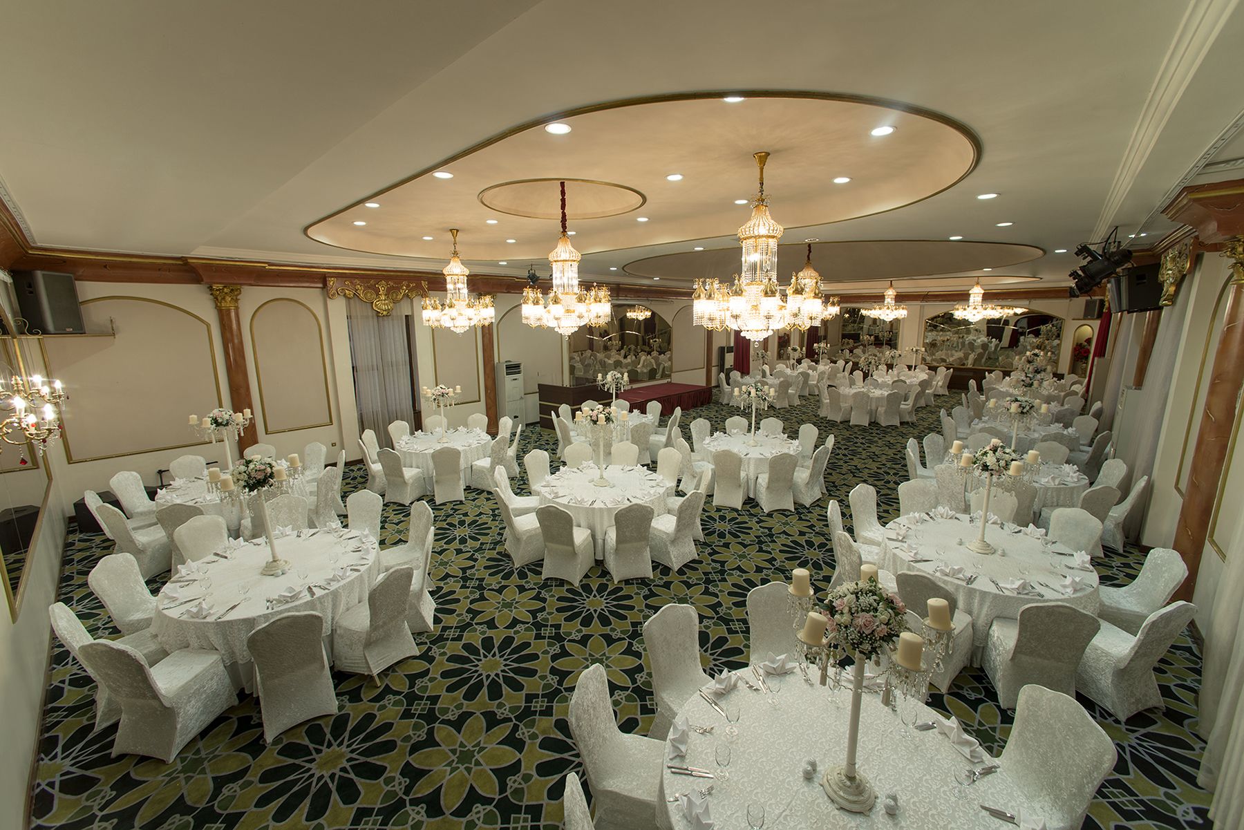 Un salón de banquetes con mesas redondas, sillas blancas, candelabros y una alfombra estampada.