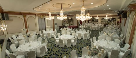 Un salón de banquetes con mesas redondas, sillas blancas, candelabros y una alfombra estampada.