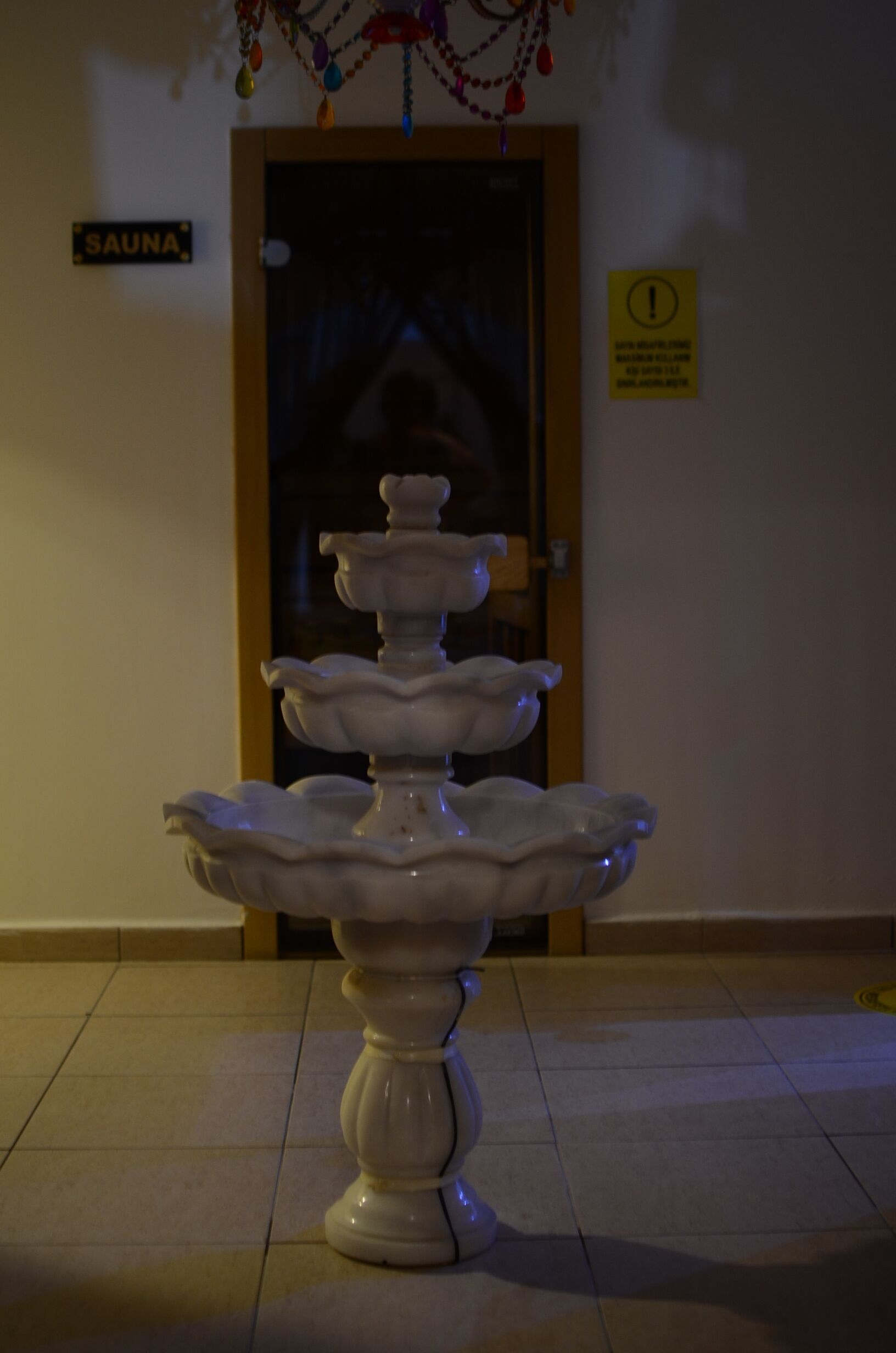 A white, multi-tiered fountain in a lobby with a sign reading "SAUNA" above a door.