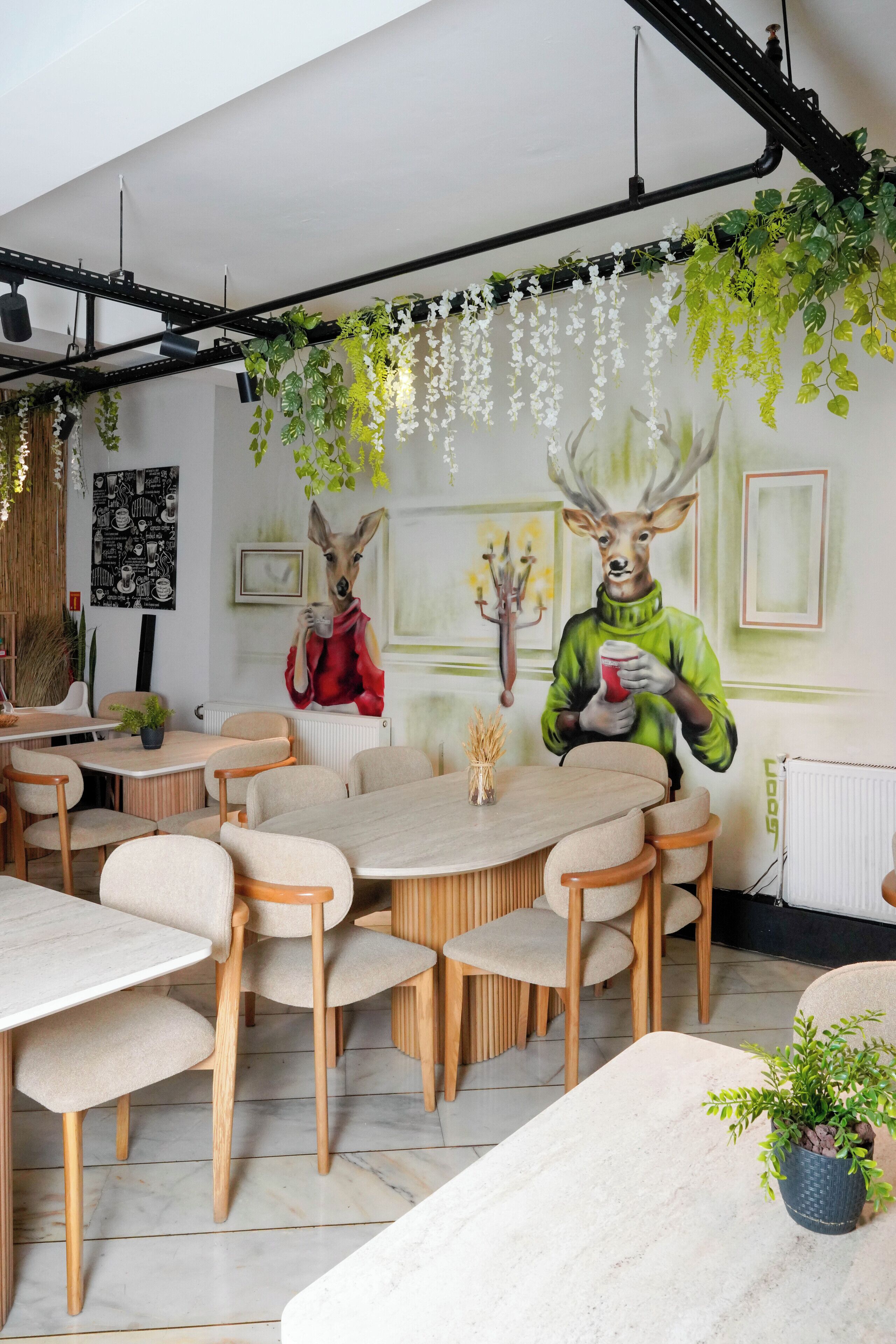 A dining area with round tables and chairs, a mural with deer figures, and hanging plants.