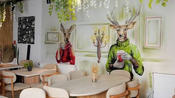 Comedor con mesas y sillas redondas, un mural con figuras de ciervos y plantas colgantes.
