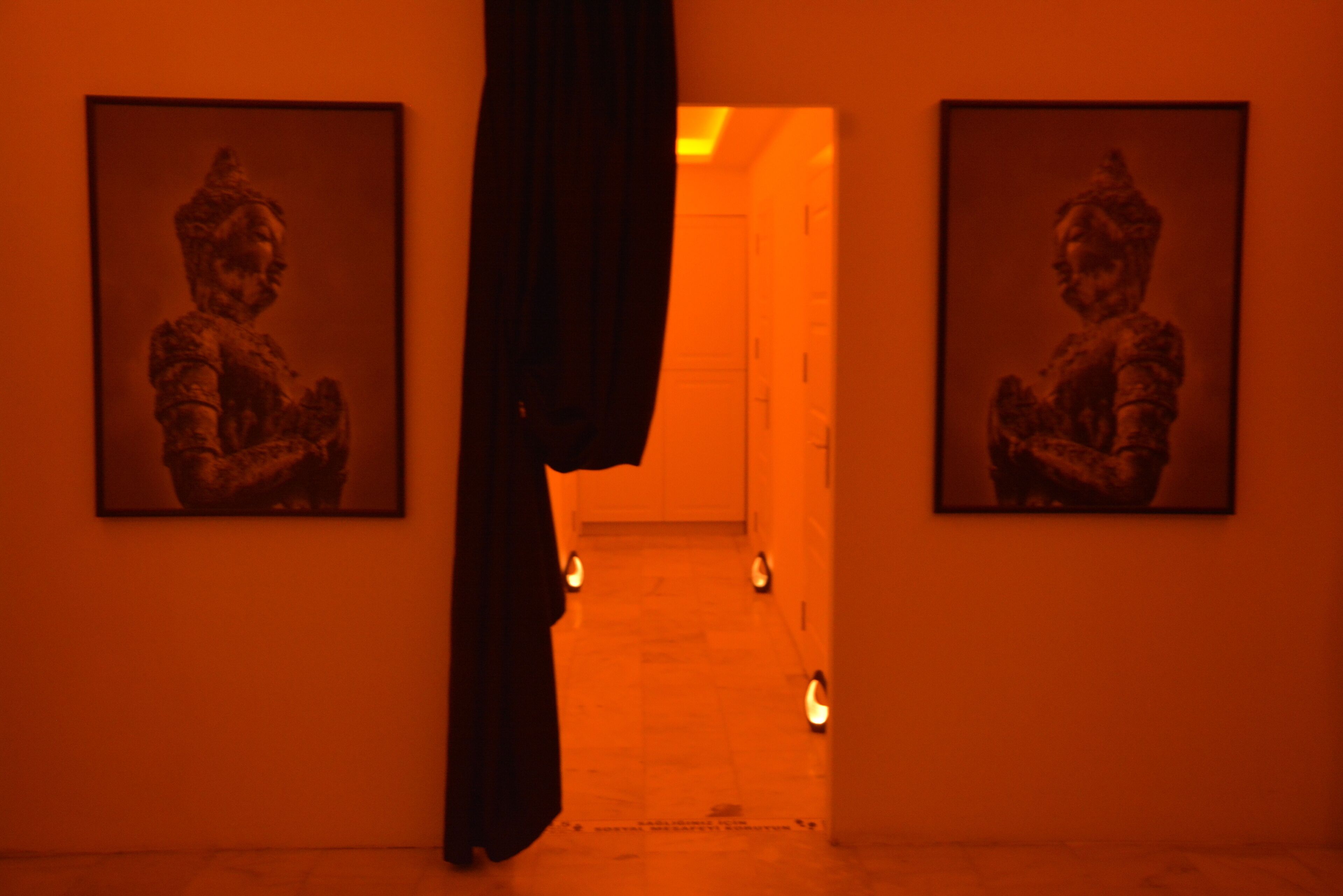 A corridor with two framed artworks on the walls, a dark curtain partially covering the entrance, and a series of small lights on the floor leading to a door at the end.