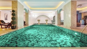 Une vaste piscine intérieure, dont le sol et les murs sont recouverts de carreaux, entourée de colonnes de marbre et surmontée d’un plafond doté d’un éclairage encastré.
