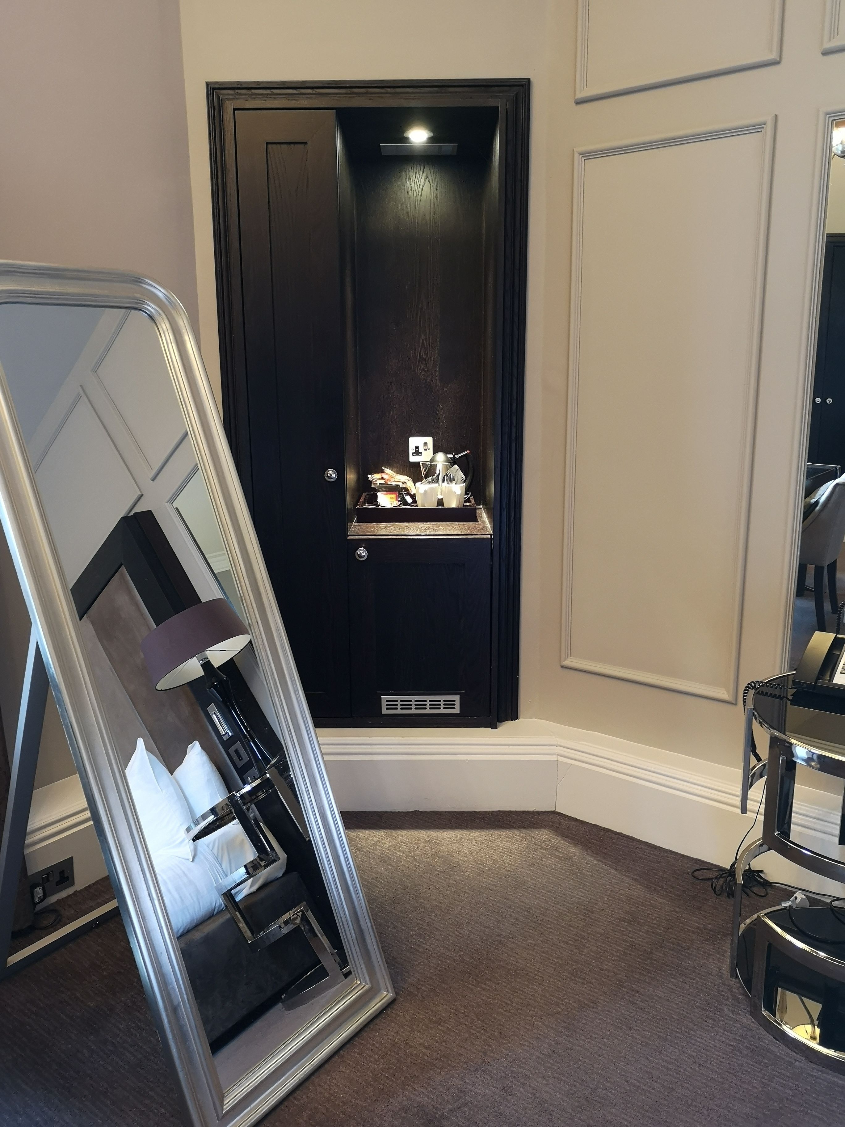A hotel room with a large mirror, a black door, a small table with a lamp, and a piano.