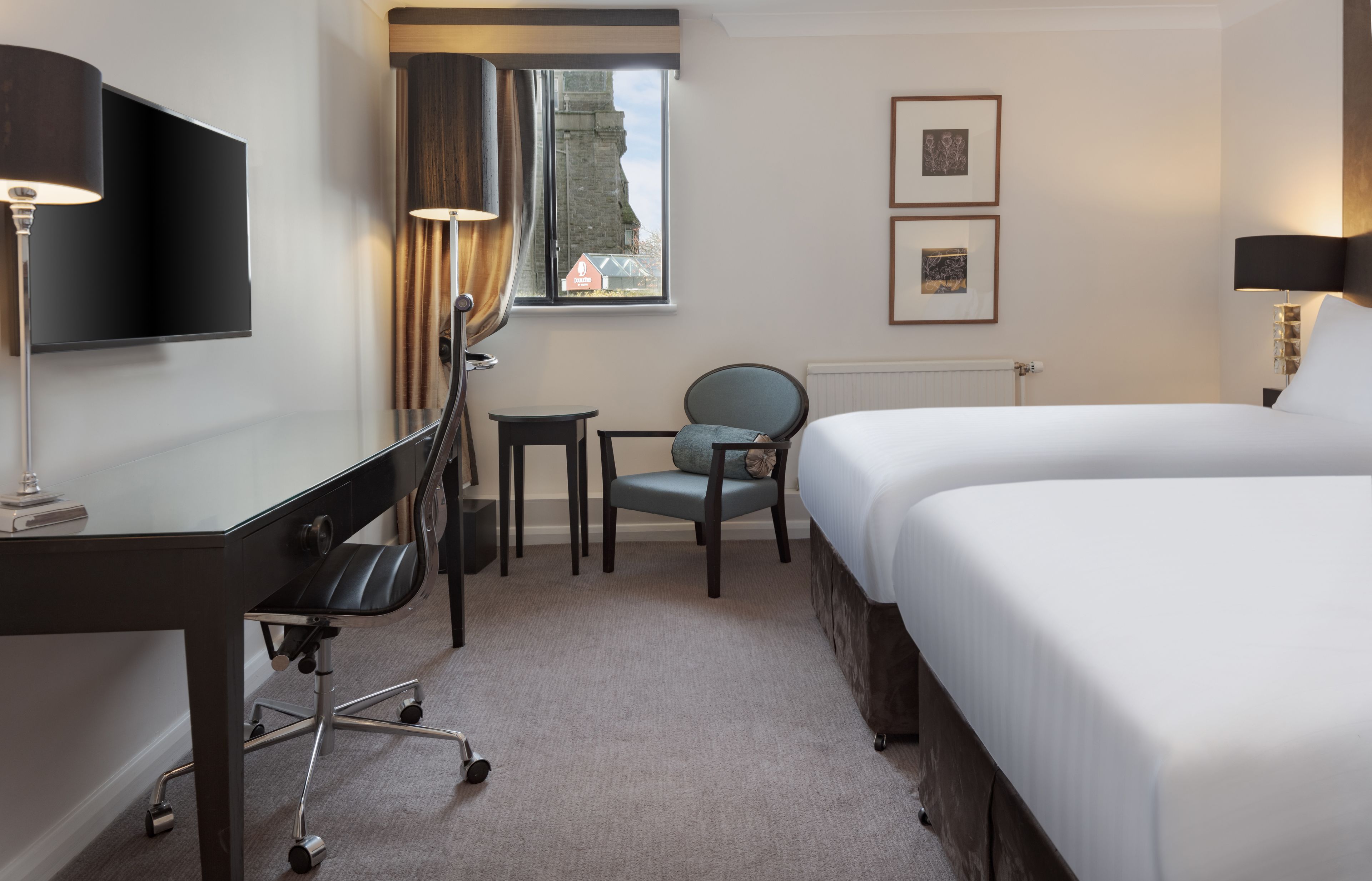 Habitación de hotel con cama, escritorio, silla, televisor y ventana con cortinas.