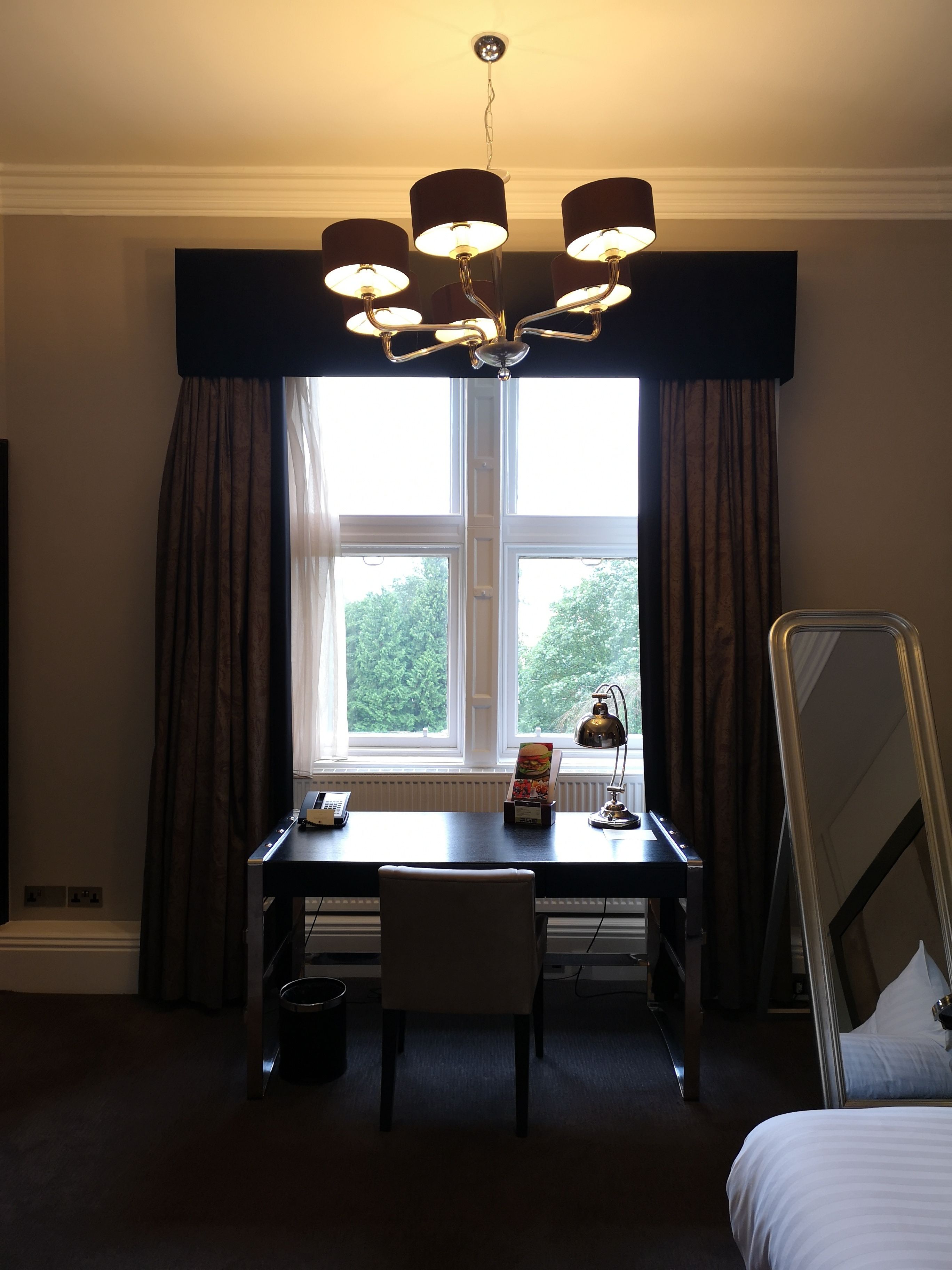 Une chambre d’hôtel équipée d’un bureau, d’une chaise et d’un lit. On y trouve une fenêtre avec des rideaux, une lampe et une table de chevet avec une lampe.