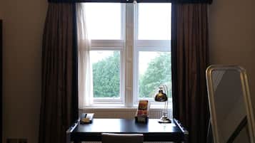 Habitación de hotel con escritorio, silla y cama. Se ve una ventana con cortinas, una lámpara y una mesita de noche con lámpara.