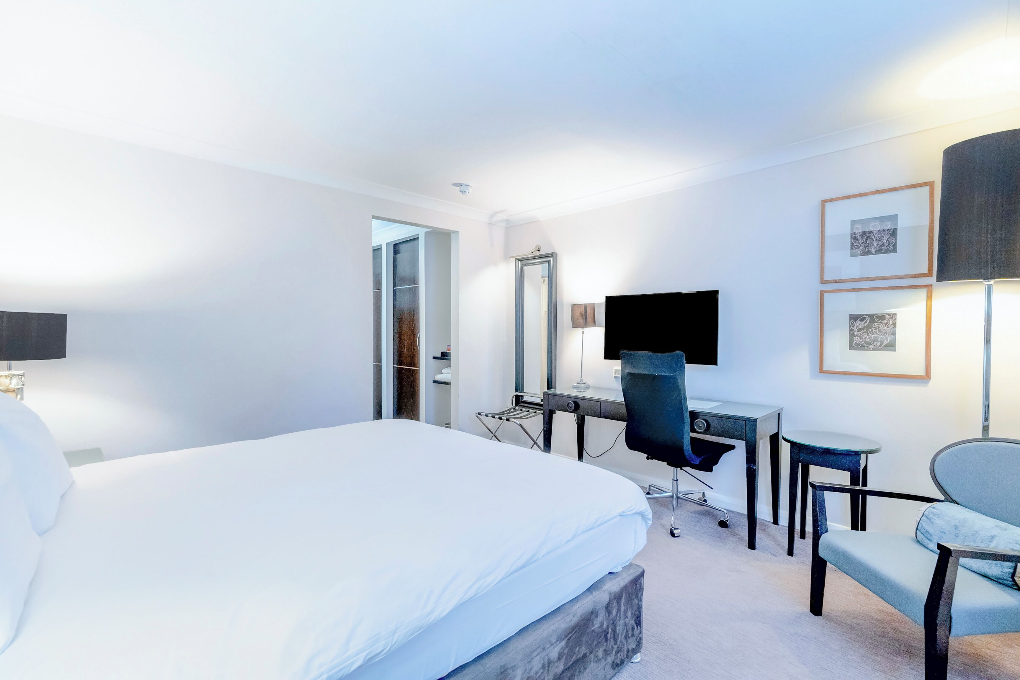 A hotel room with a large bed, a desk with a chair, a TV, and framed pictures on the wall.
