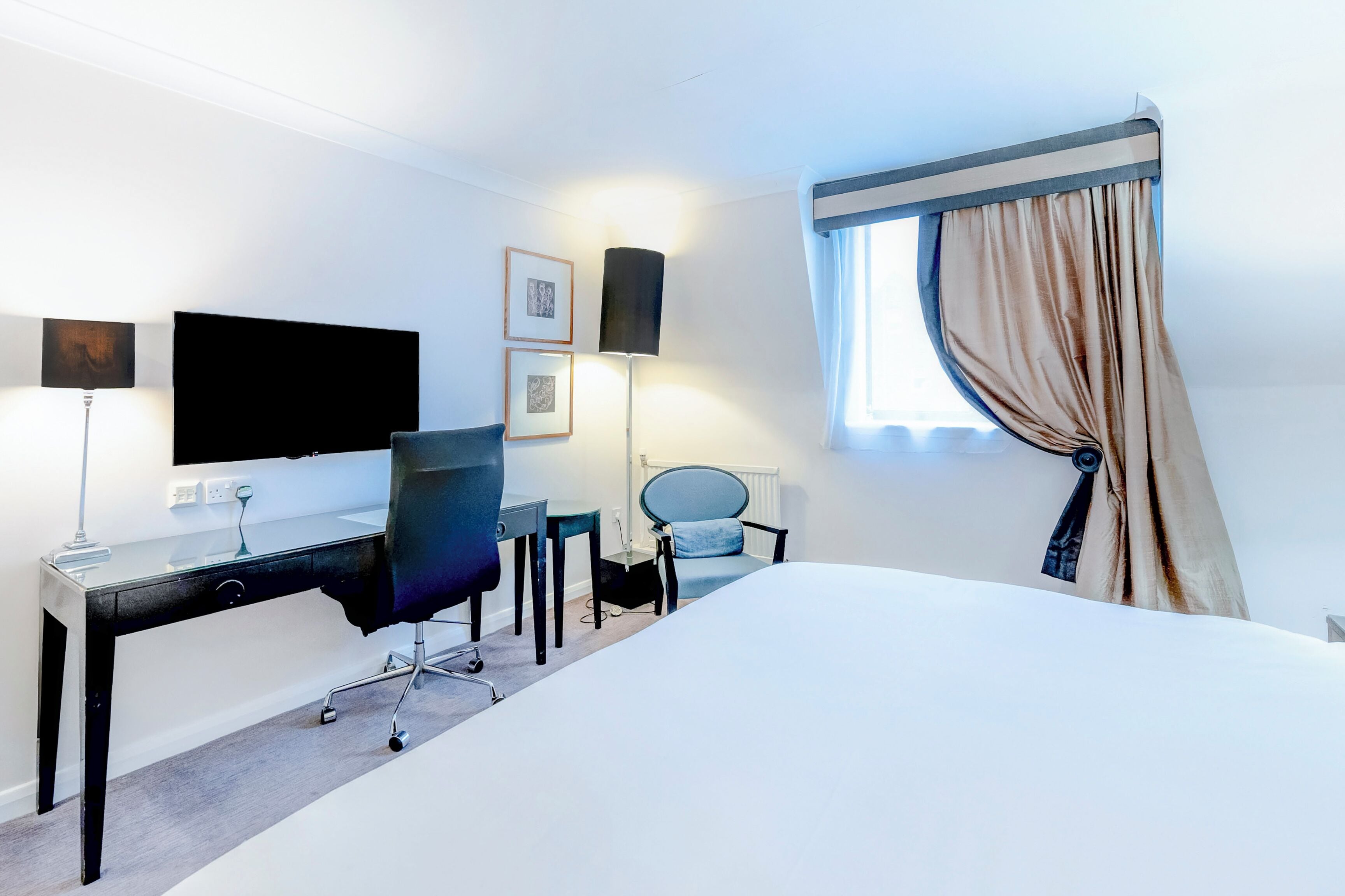 A hotel room with a bed, a desk with a chair, a flat-screen TV, a lamp, and a window with curtains.