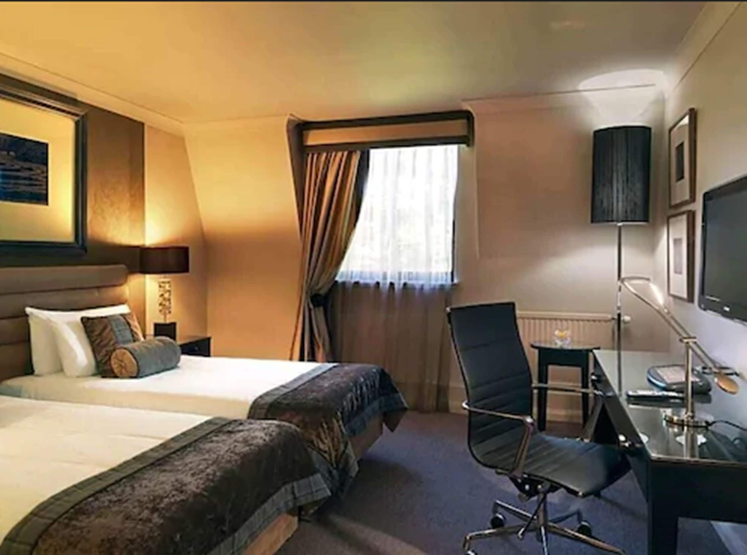 A hotel room with two beds, a desk, a chair, a TV, and a window with curtains.