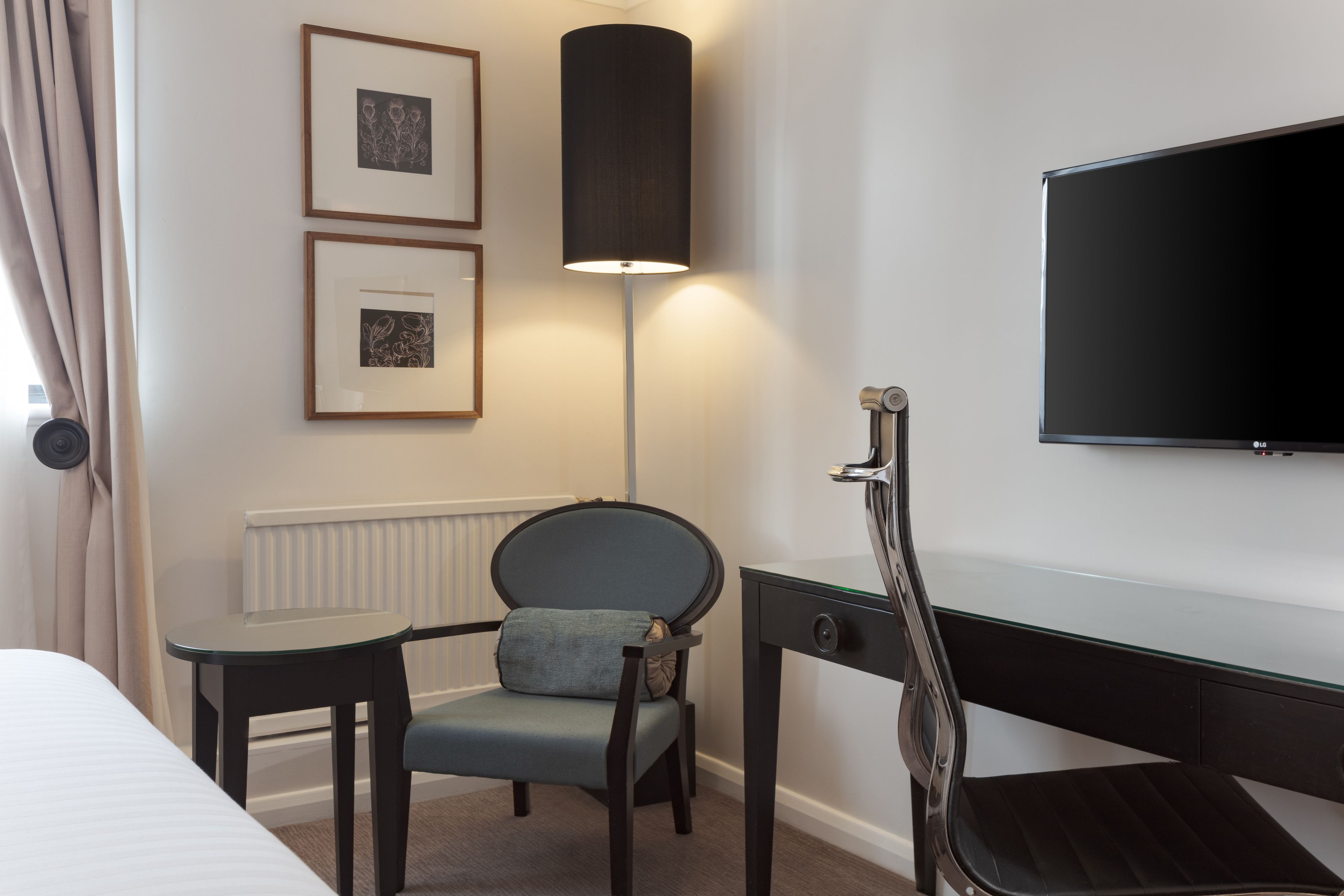 Habitación de hotel con cama, escritorio con silla, lámpara de pie y televisor de pantalla plana en la pared.