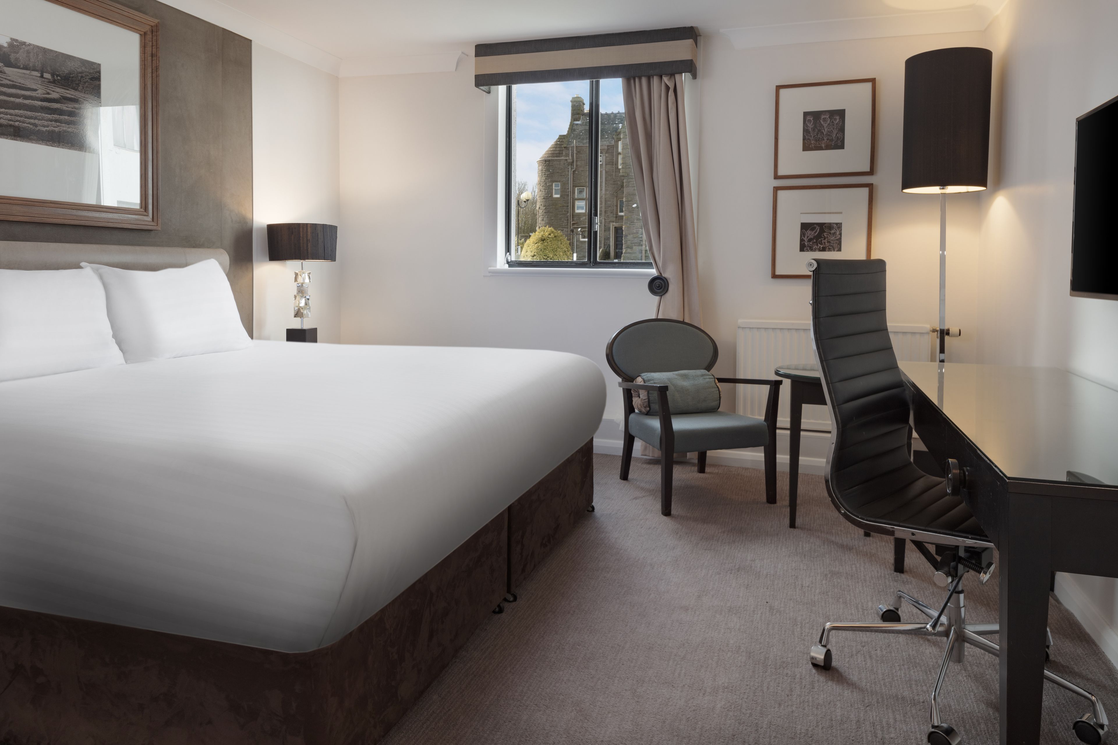 A hotel room with a large bed, a desk with a chair, a small table, and a window with curtains.