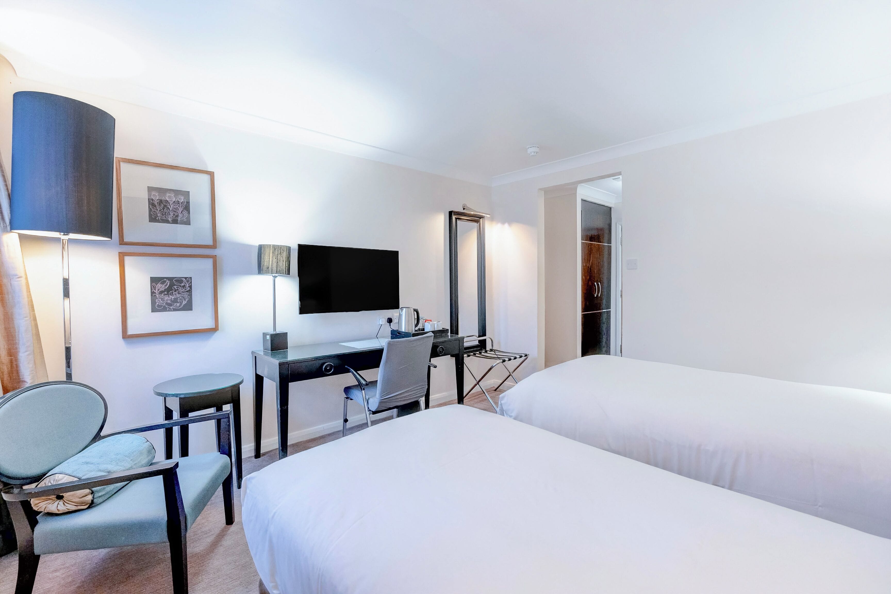 Ein Hotelzimmer mit zwei Betten, einem Schreibtisch mit Computer, einem Sessel, einem Fernseher und gerahmten Bildern an der Wand.