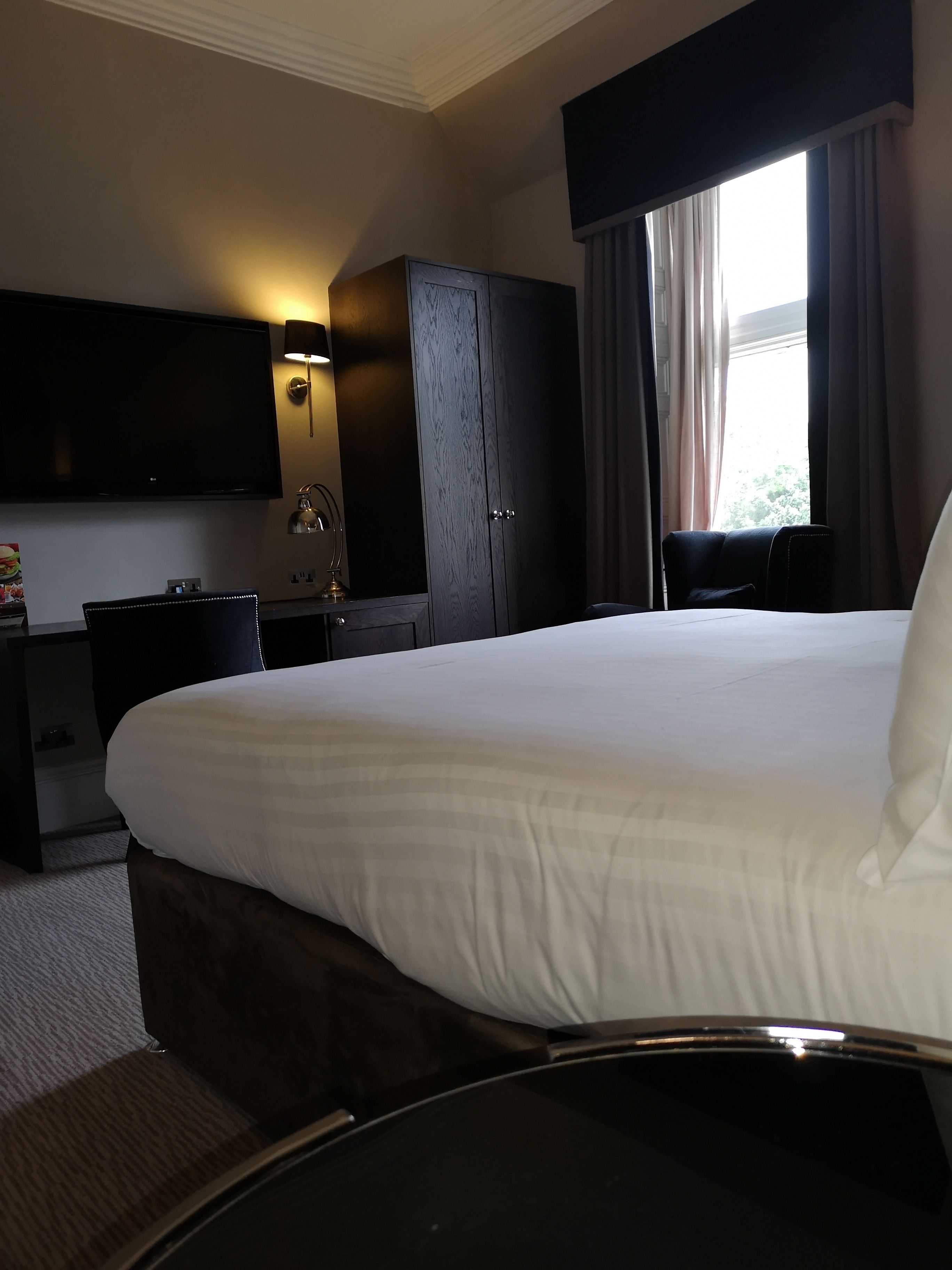 A hotel room with a large bed, a flat-screen TV mounted on the wall, a desk with a lamp, a wardrobe, and a window with curtains.
