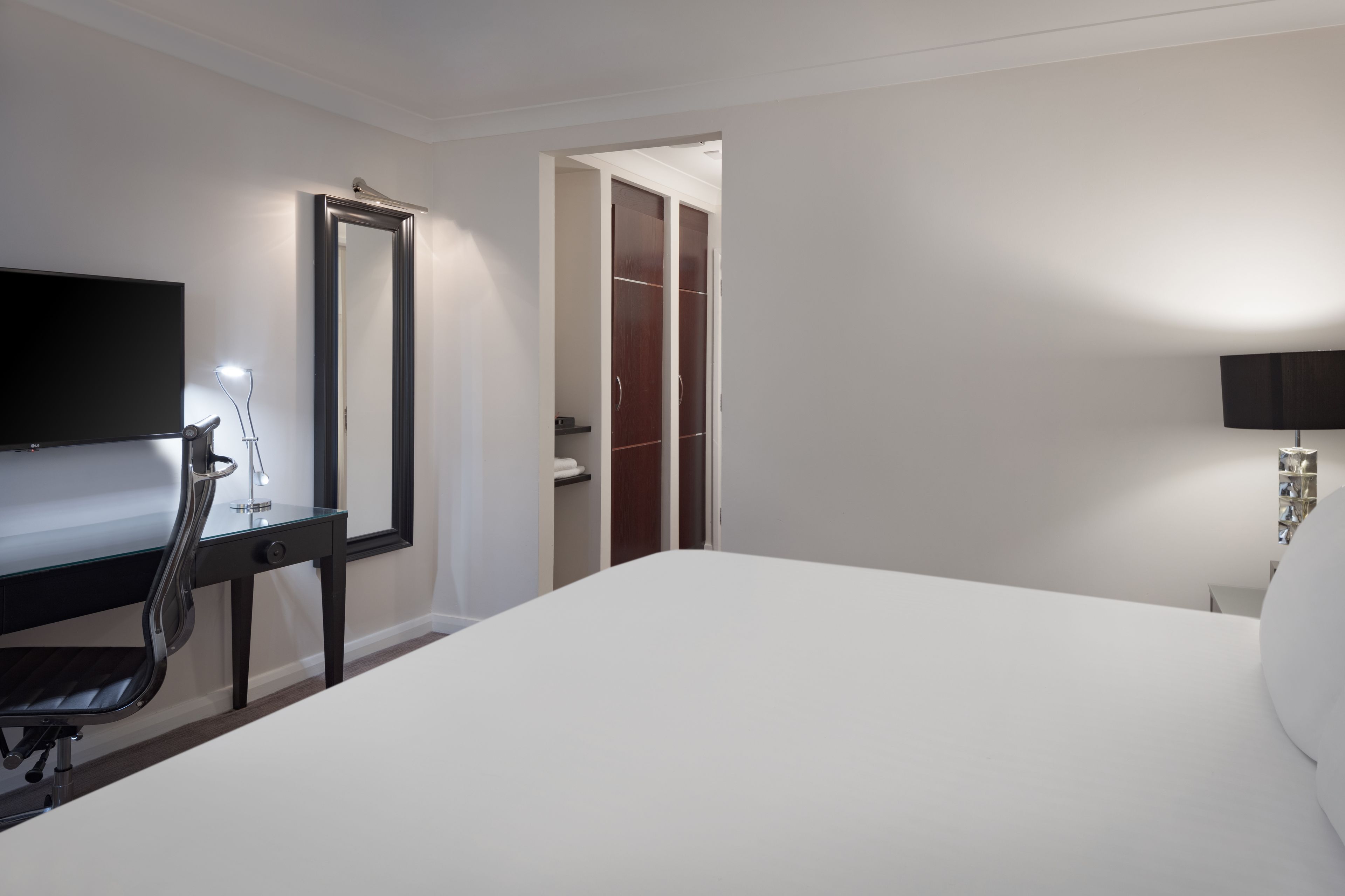 A hotel room with a bed, a desk with a chair, a TV, a mirror, a wardrobe, and a bedside lamp.