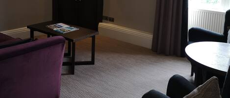 Habitación de hotel con un sofá de color oscuro, una mesita con revistas, un televisor montado en la pared y una ventana con cortinas.