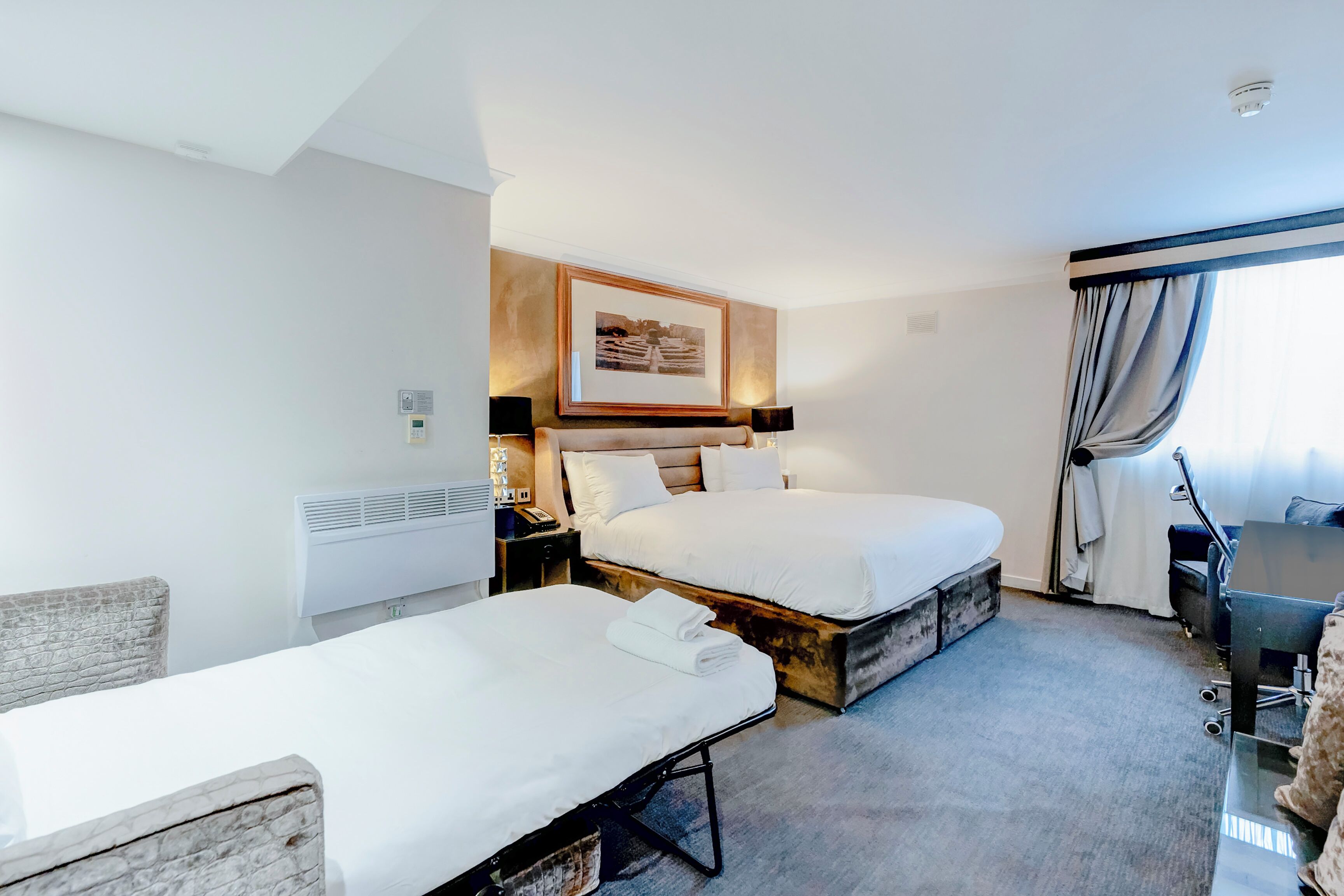 Une chambre d’hôtel avec deux lits simples, un bureau, une chaise, une grande fenêtre avec des rideaux et un tableau encadré au mur.