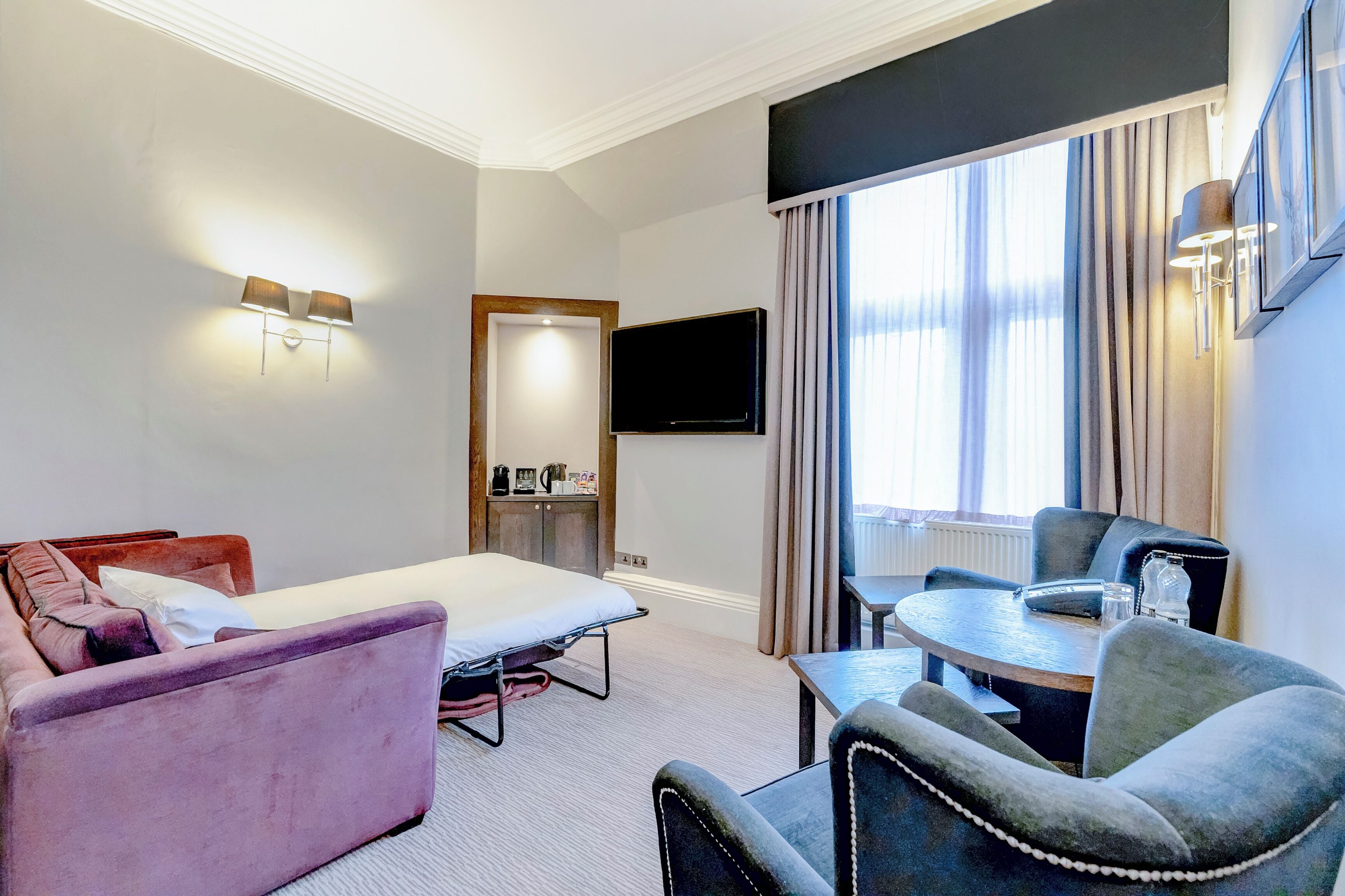 A hotel room with a bed, a sofa, a dining table with chairs, a TV mounted on the wall, and a window with curtains.