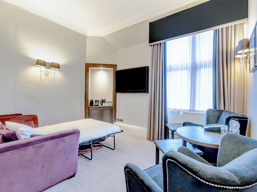 A hotel room with a bed, a sofa, a dining table with chairs, a TV mounted on the wall, and a window with curtains.