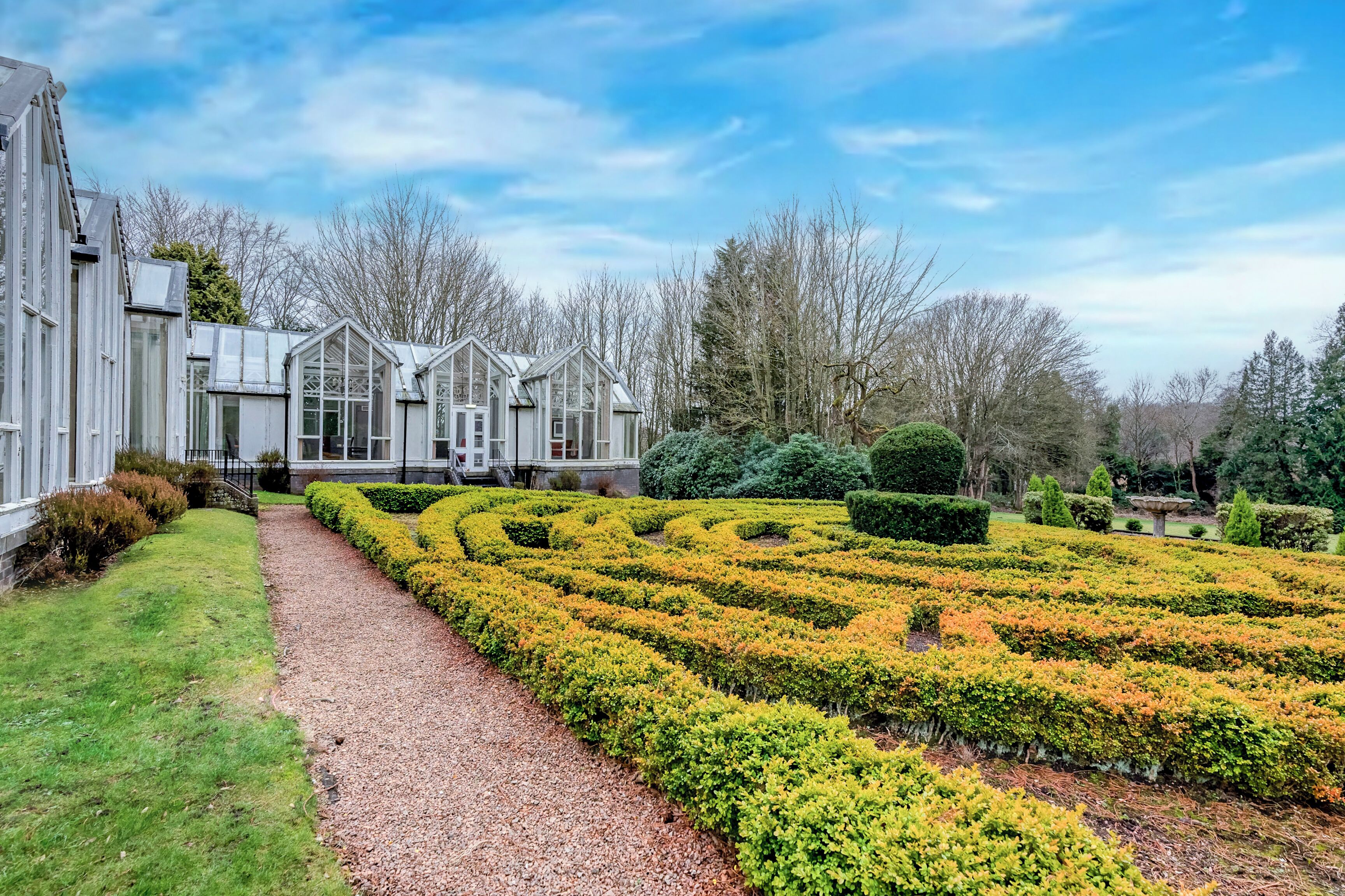 Un jardin à la française, avec un chemin de gravier, des haies taillées avec soin et un grand bâtiment doté de nombreuses fenêtres et d’un toit à pignon.