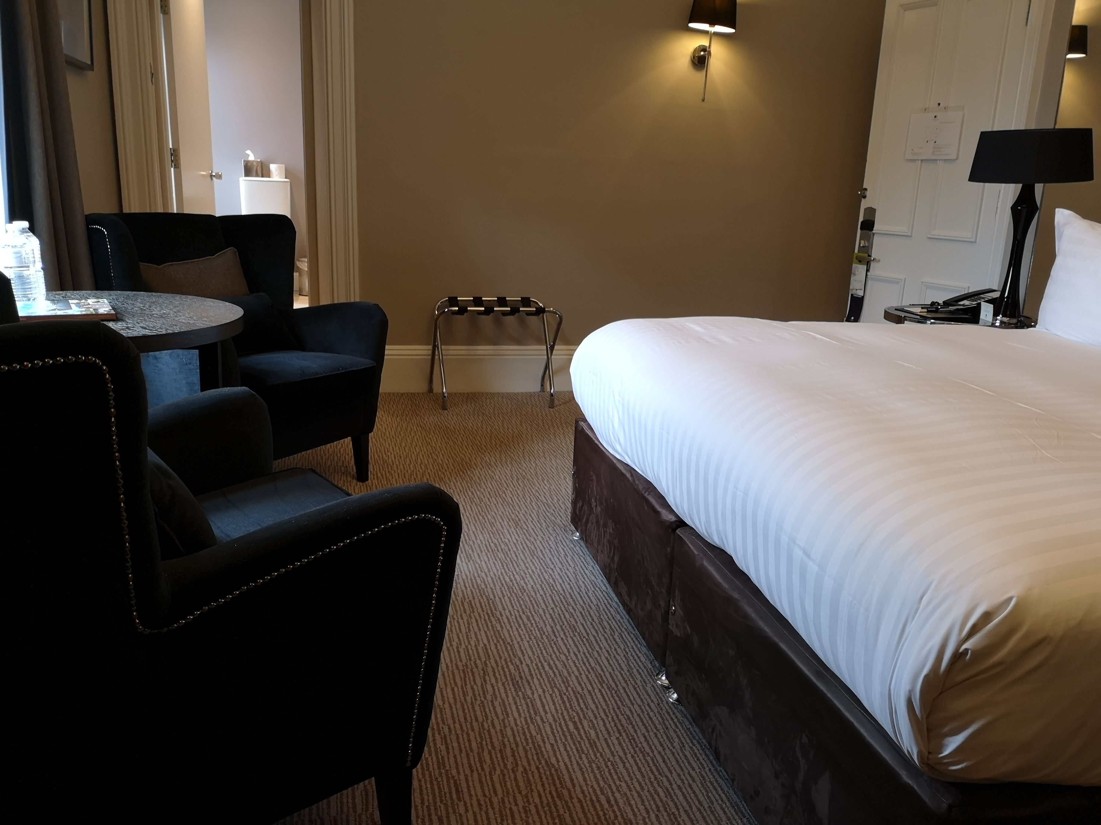 A hotel room with a large bed, a desk with a chair, a small table, and a standing lamp.