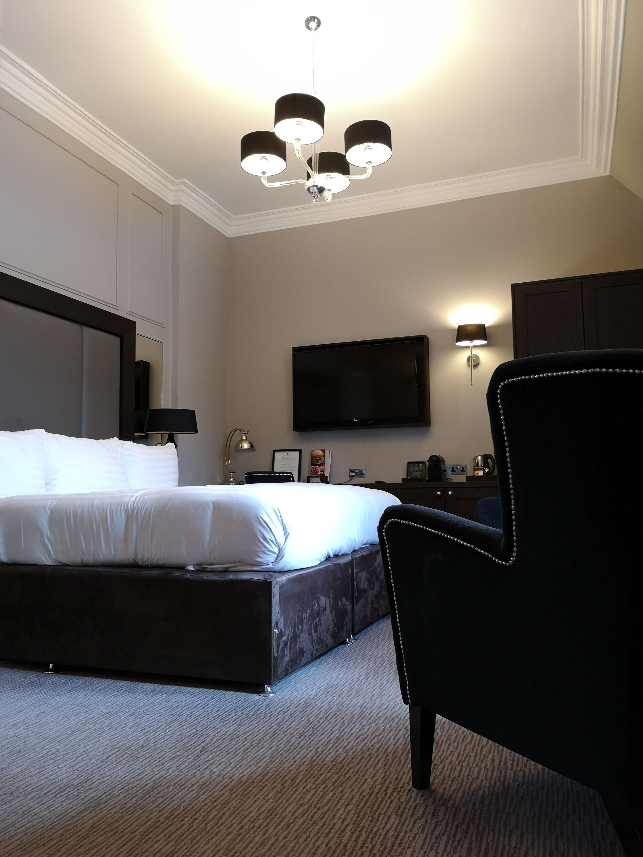 Habitación de hotel con una cama grande, un televisor de pantalla plana montado en la pared, un escritorio con lámpara y una silla con detalles perlados.